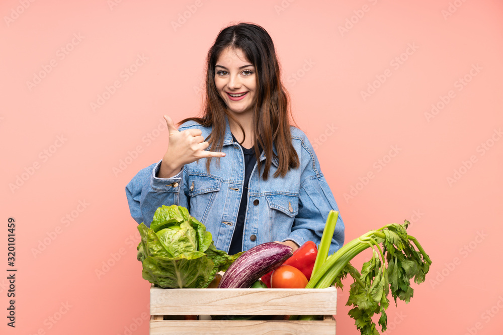 Young farmer woman holding a basket full of fresh vegetables making phone gesture