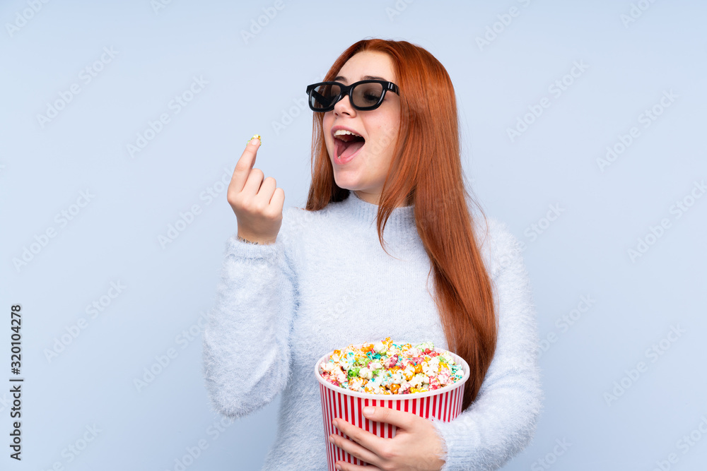 Redhead teenager girl over isolated blue background with 3d glasses and holding a big bucket of popcorns