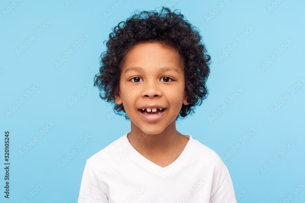 Closeup portrait of amazing cheerful little boy with curly hairdo in white T-shirt looking at camera with happy carefree smile and missed milk teeth. indoor studio shot isolated on blue background - obrazy, fototapety, plakaty 