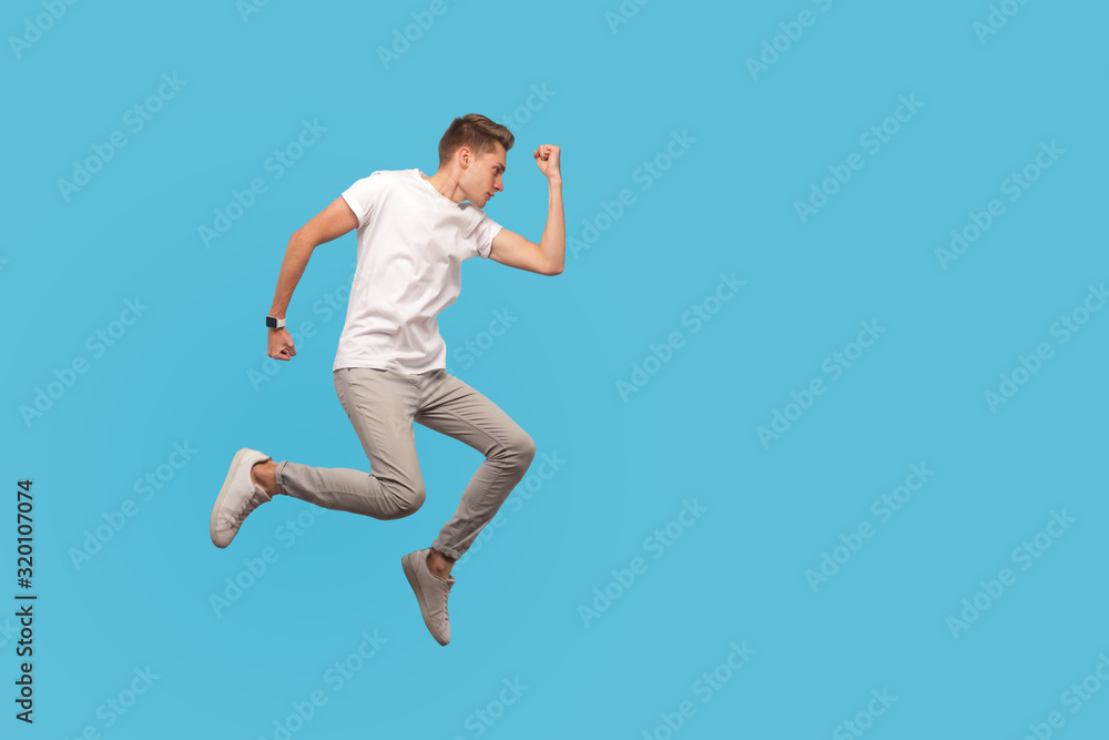 Full length, motivated man in white t-shirt and casual pants running in air with serious confident determined expression, hurrying to meet dream, goal. indoor studio shot isolated on blue background