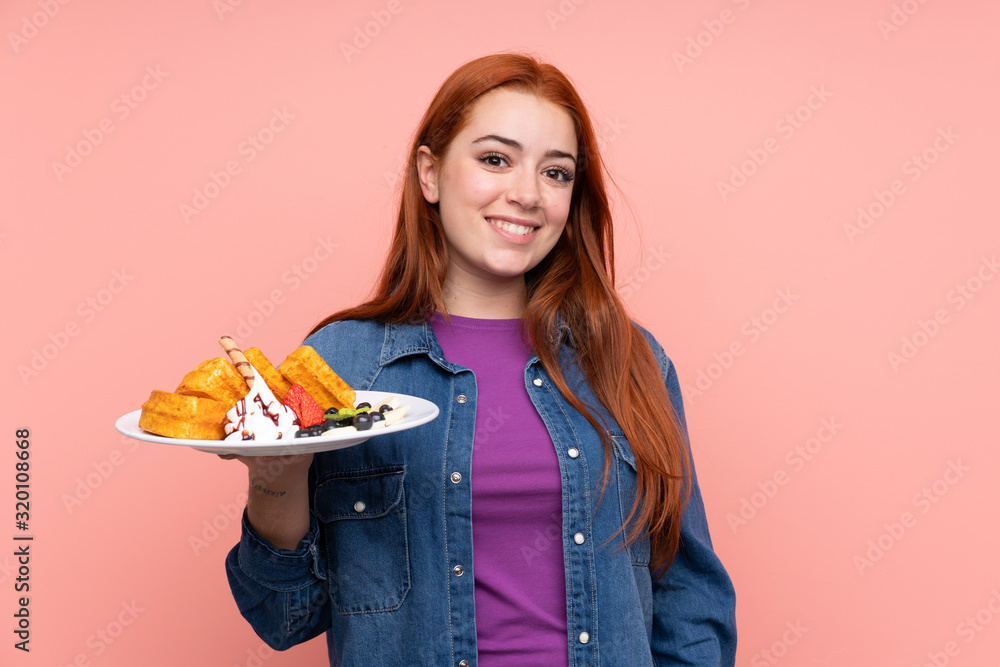 Redhead teenager girl holding waffles over isolated pink background smiling a lot