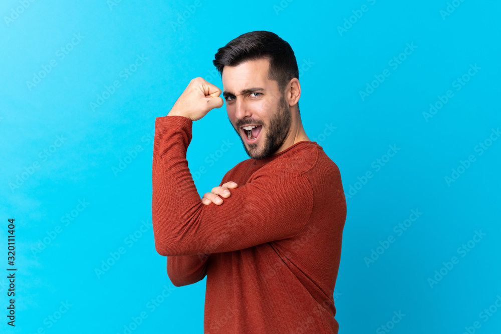 Young handsome man with beard over isolated blue background making strong gesture Young handsome man with beard over isolated blue background making strong gesture