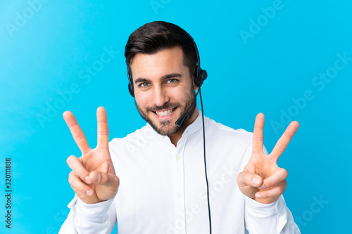 Telemarketer man working with a headset over isolated blue background smiling and showing victory sign © luismolinero