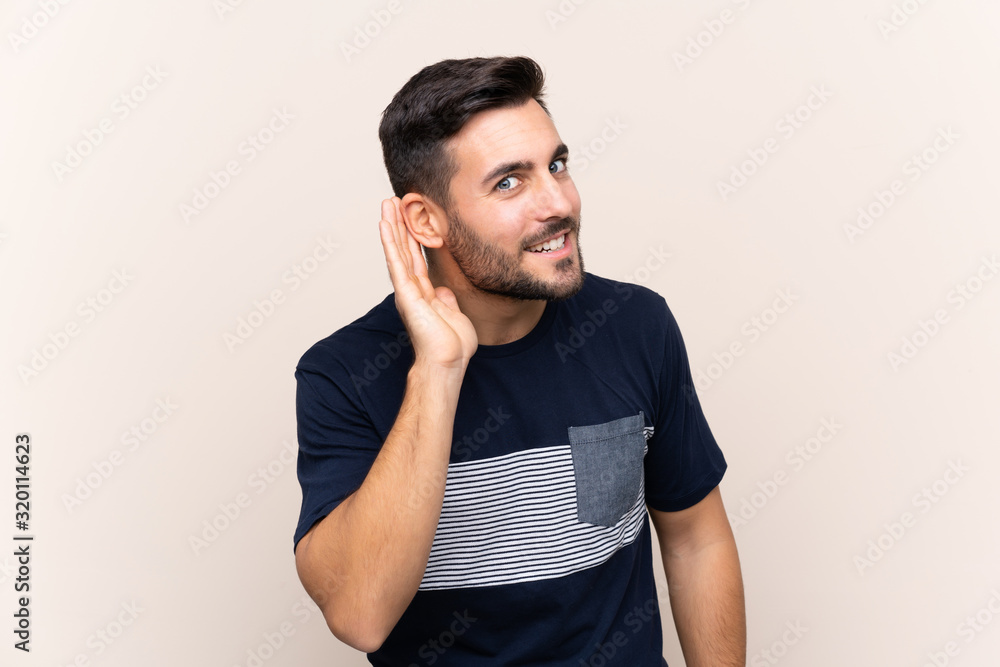 Young handsome man with beard over isolated background listening to something by putting hand on the ear Young handsome man with beard over isolated background listening to something by putting hand on the ear