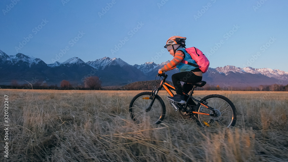 One caucasian children rides bike in wheat field. Little girl riding black orange cycle on background of beautiful snowy mountains. Biker motion ride with backpack and helmet. Mountain bike hardtail.