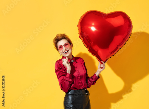 Happy woman holds red heart shape balloon. Photo of elderly woman in love in red shirt on yellow background