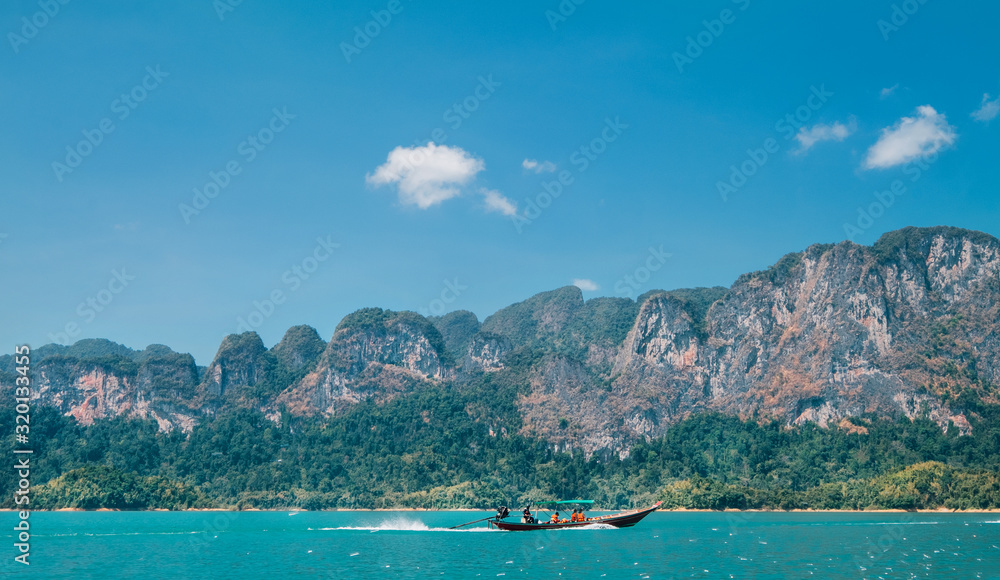 Wooden motorboat vessel using a long propeller shaft sailing with passengers on Thai Khao Sok Lake with foresty mountains around. Exotic countries traveling concept image.