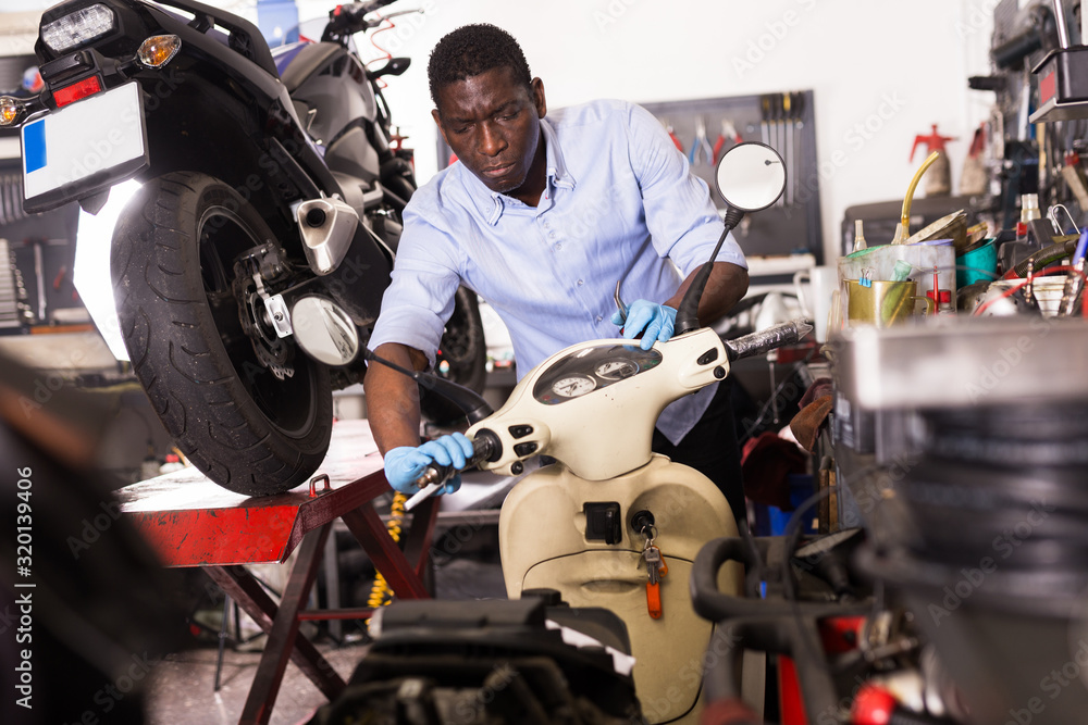 Fototapeta Male afro american worker repairing scooter in motorcycle workshop