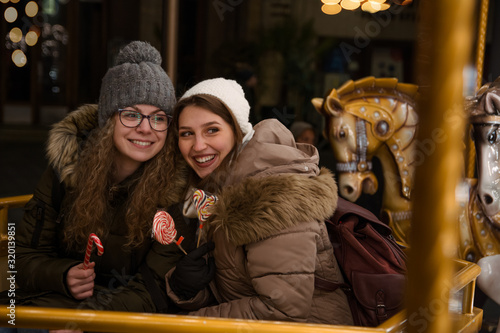 Two female friends riding on the carousel with horses at the winter festival