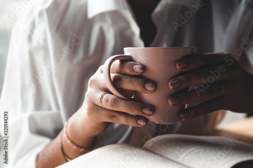Woman in the morning drinks coffee and reads old book in a white shirt. Education, drink. manicure
