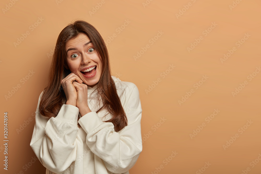 Joyful lovely woman fools around with friends, has positive gaze at camera, keeps hands near face, tilts head, wears casual white hoodie, opens mouth, poses against brown background, blank space Joyful lovely woman fools around with friends, has positive gaze at camera, keeps hands near face, tilts head, wears casual white hoodie, opens mouth, poses against brown background, blank space