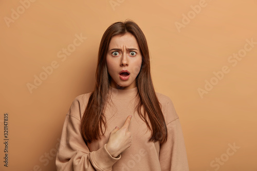Fotografi Stunned brunette European woman indicates at herself with indignant stupefied expression, shocked being chosen, frowns eyebrows, wears loose sweatshirt, models over beige wall