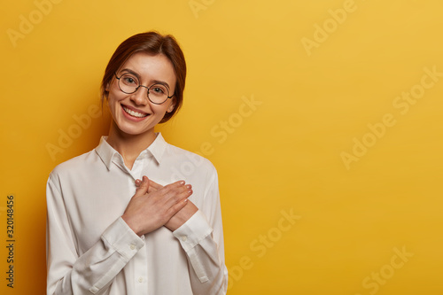 Charming positive European woman presses hands to chest, expresses gratitude for gift, appreciates help, wears round glasses and white shirt, stands against yellow background, looks with admiration photo
