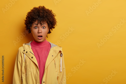 Concerned frightened curly adult woman feels fear and panic because of terrible accident, keeps mouth opened, dressed in casual anorak, gasps amazed, isolated on yellow wall, copy space area