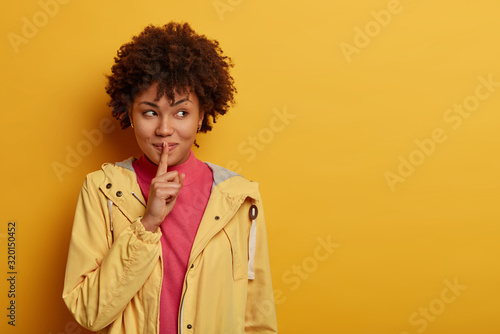 Portrait of attractive mysterious woman tells pleasant secret to close friend looks aside shows shh gesture, looks with positive expression away, presses index finger on mouth, prepares surprise gift