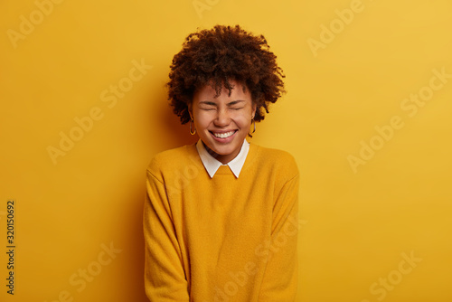 Photo of joyful emotional dark skinned woman squints face, smiles broadly, feels amused and entertained during party, wears casual jumper, models over bright yellow background. Emotions concept © wayhome.studio 