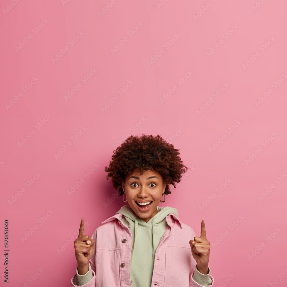 Glad curly haired woman tells about new product, indicates upwards on copy space, has positive toothy smile, suggests amazing cool offer, poses against pink wall. Follow this way. Advertisement