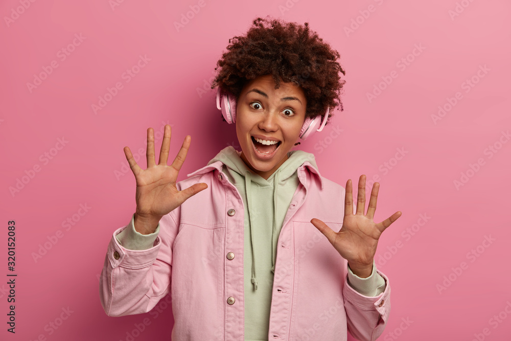 Joyful cheerful girl raises palms, has funny expression, tilts head and laughs, listens music in headphones, enjoys favourite track, wears fashionable clothes, stands indoor against rosy wall Joyful cheerful girl raises palms, has funny expression, tilts head and laughs, listens music in headphones, enjoys favourite track, wears fashionable clothes, stands indoor against rosy wall