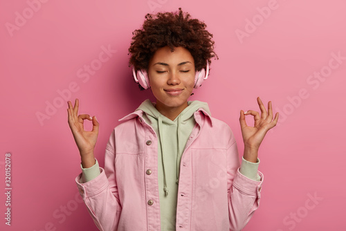 Relaxed beautiful woman makes zen gesture, meditates while listens peaceful music in headphones, has relaxation session during spare time, closes eyes, wears stereo headphones and sweatshirt