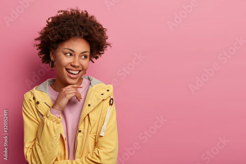 Millennial girl looks joyfully aside, keeps hand under chin, has casual talk with friend, smiles gently, discuss nice ideas for personal business, wears casual windbreaker, isolated on pink background