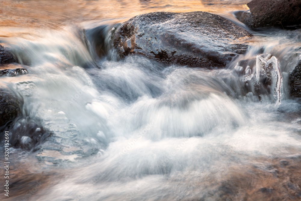 Dark, frozen stone and branch covered by ice in the middle of cold mountain creek during winter, long exposure blurred motion water, lighten by golden sunlight Dark, frozen stone and branch covered by ice in the middle of cold mountain creek during winter, long exposure blurred motion water, lighten by golden sunlight