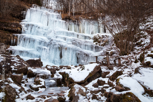 Beautiful, scenic, frozen Tupavica waterfall with hanging icicles and mountain creek with orange rocks and wooden bench and picnic table