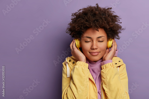 Horizontal shot of lovely woman meloman uses electronic headphones, feels enjoyment and pleasure from popular sound track, closes eyes, wears yellow anorak, isolated on purple wall, copy space
