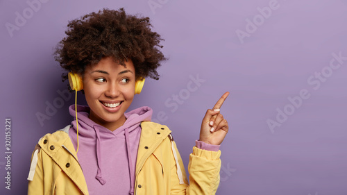 Pretty dark skinned woman with Afro hairstyle, points index finger on blank space, shows promo with glad expression, wears stereo headphones on ears, purple hoodie, has good mood. People, lifestyle