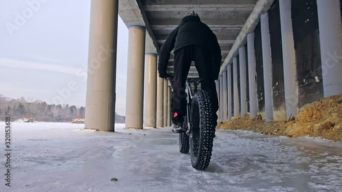 Professional extreme sportsman biker riding fat bike in outdoors. Cyclist ride in winter on snow ice under bridge. Man do trick on mountain bicycle with big tire in helmet glasses. Slow motion 180fps. photo