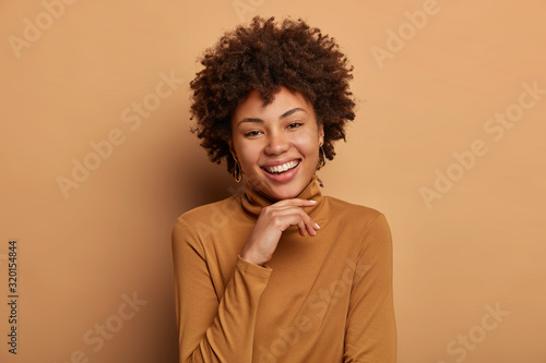 Portrait of cute African American woman gazes with tenderness and delight, keeps hands under chin, laughs at positive funny story wears casual brown turtleneck models indoor recalls something pleasant