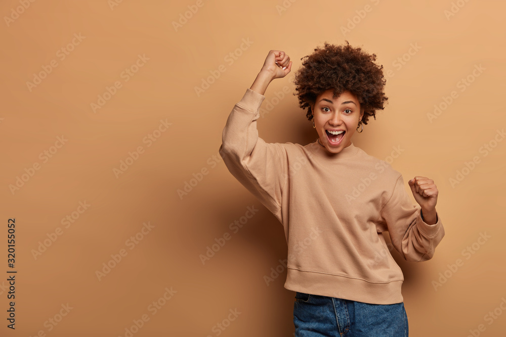 Triumphing joyful woman celebrates win, feels lucky and upbeat, yells yes and dances happily, makes fist pump, cheers something, wears brown sweater and jeans, laughs loudly. Copy space on brown wall Triumphing joyful woman celebrates win, feels lucky and upbeat, yells yes and dances happily, makes fist pump, cheers something, wears brown sweater and jeans, laughs loudly. Copy space on brown wall