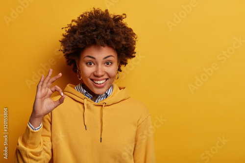 Not bad. Glad hipster girl shows okay gesture, approval sign, accepts good idea, smiles positively, wears yellow hoodie, gives approval, takes everything under control, says yes to new opportunities photo
