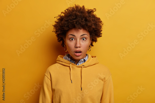 Speechless impressed curly haired teenager gasps from wonder, holds breath, wears yellow hoodie, feels surprisement and astonishment, isolated over vivid background. People and emotions concept © wayhome.studio 