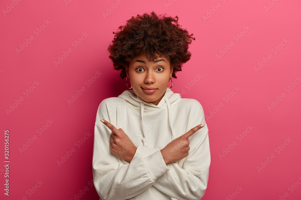 Photo of confused dark skinned woman crosses hands and points at different sides, shows two directions, chooses best option, wears white sweatshirt, isolated on pink background. Difficult choice Photo of confused dark skinned woman crosses hands and points at different sides, shows two directions, chooses best option, wears white sweatshirt, isolated on pink background. Difficult choice