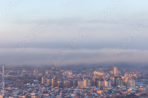 Moody, misty cityscape with buildings and houses lighten by golden sunrise sun and rooftops covered by first snow © Nikola