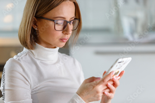 Serious businesswoman wears white turtleneck sweater and glasses using modern smartphone in marble case, browse internet, buying goods online, blurred background. Social networks addiction, nomophobia