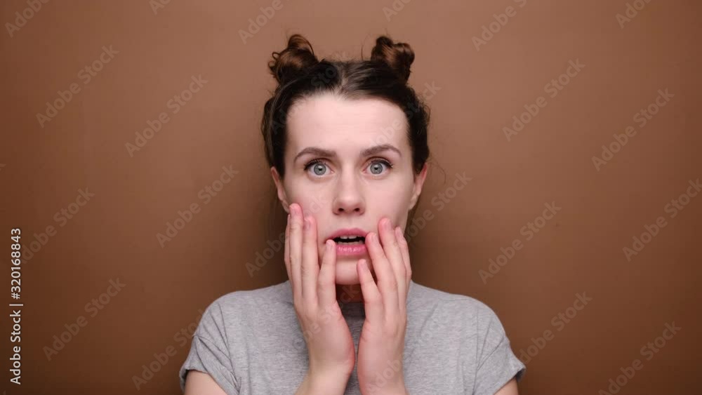 Bewildered young brunette woman touches cheek, opens mouth, has eyes full of fear and disappointment, wears grey t-shirt, isolated over brown background. Human facial expressions Bewildered young brunette woman touches cheek, opens mouth, has eyes full of fear and disappointment, wears grey t-shirt, isolated over brown background. Human facial expressions