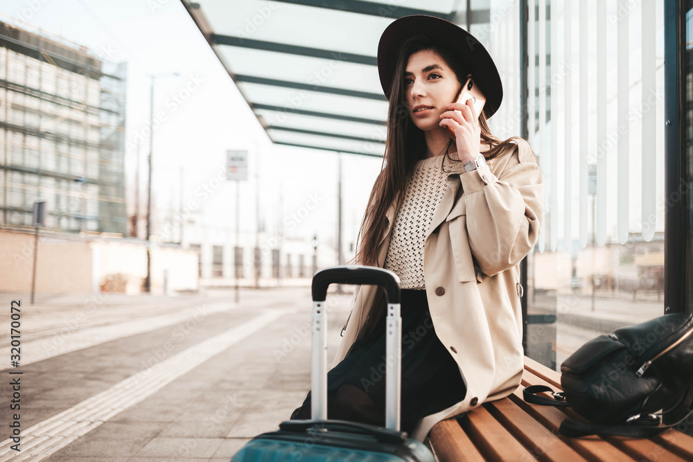 Fototapeta premium Young beautiful traveler girl is waiting at the public transport stop talking on the smart phone