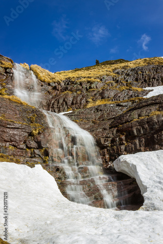 Large  scenic  beautiful waterfall water falling from red rocky cliff under the snow during transition period between winter and spring