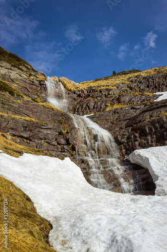 Large, scenic, beautiful waterfall water falling from red rocky cliff under the snow during transition period between winter and spring