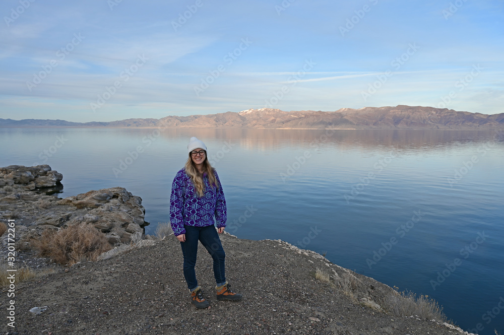 Young woman in blue sweater enjoys view of tranquil Pyramid Lake, Nevada from rock formation on coast on a calm clear winter afternoon.