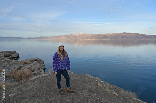 Young woman in blue sweater enjoys view of tranquil Pyramid Lake, Nevada from rock formation on coast on a calm clear winter afternoon.