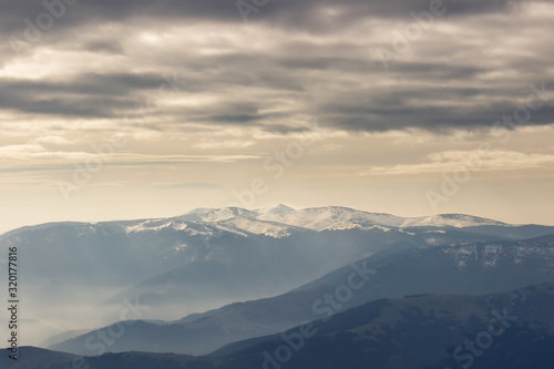 Moody, dramatic sky above pointy, distant, impressive Kom summit in Bulgaria covered by snow and misty, foreground mountain layers © Nikola