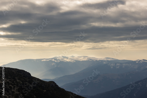 Fototapeta Naklejka Na Ścianę i Meble -  Moody, dramatic sky above pointy, distant, impressive Kom summit in Bulgaria covered by snow and misty, foreground mountain layers