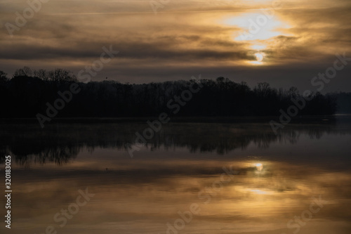 The orange glow of the morning sun is peaking from behind the clouds. The light and shadows of the trees reflect off the still calm water.