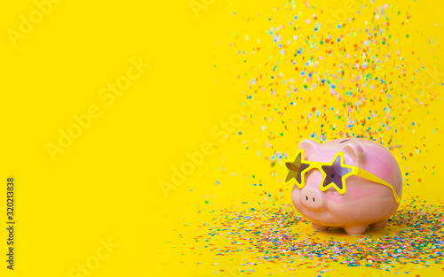 Fotografia A pink piggy bank in fun glasses at a party. Yellow background.