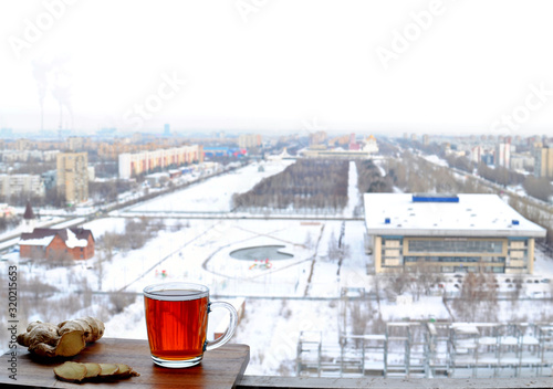 A transparent glass mug of hot black tea and sliced ​​ginger root close-up stand on a wooden surface against a blurry background of the winter panorama of the city of Togliatti.