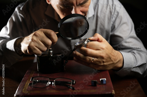 Mature man working at home as a watchmaker and jeweler composing an old clock using a screwdriver and a magnifying glass to be able to see closely