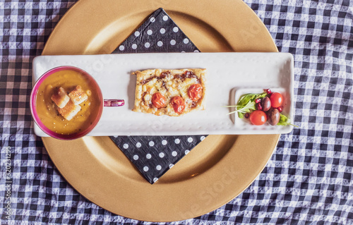 Butternut and pumpkin soup, tomato and caramalised onion pastery tart with lettuce salad served on a white rectangular plate and gold circular plate underneath photo