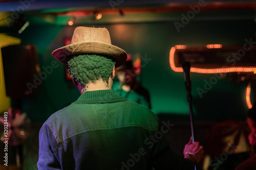 Closeup rear view of male musician wearing brown sun hat and holding microphone on stage in illuminated auditorium hall at night